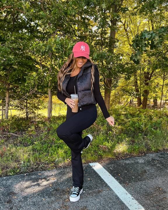 A woman wearing a black puffer vest, black leggings, sneakers, and a red baseball cap while holding an iced coffee. 