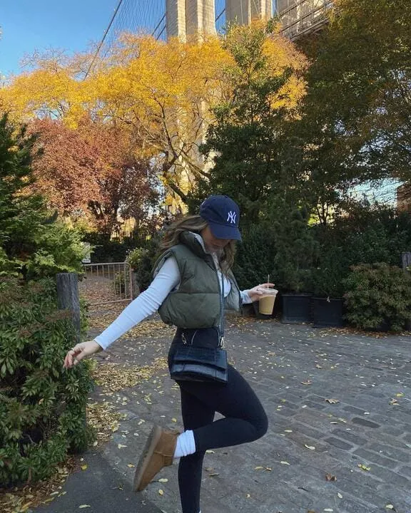 A woman wearing an olive cropped puffer vest, white long-sleeve top, black leggings, brown Ugg boots, and a navy baseball cap while holding an iced coffee.