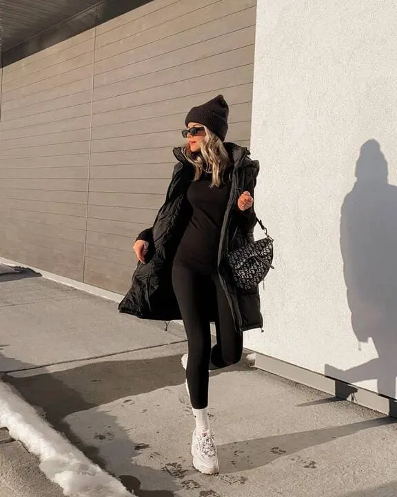 A woman wearing an all-black puffer coat, black leggings, white sneakers, a black beanie, and sunglasses while carrying a patterned handbag. 