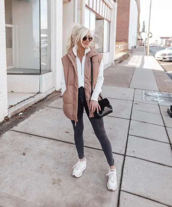 A woman wearing a beige long puffer vest, white zip-up sweater, black leggings, and chunky white sneakers while carrying a black handbag. 