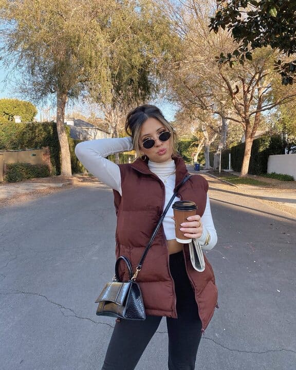 A woman wearing a brown puffer vest, white cropped turtleneck, black leggings, and sunglasses while holding a coffee cup and carrying a small black handbag. 