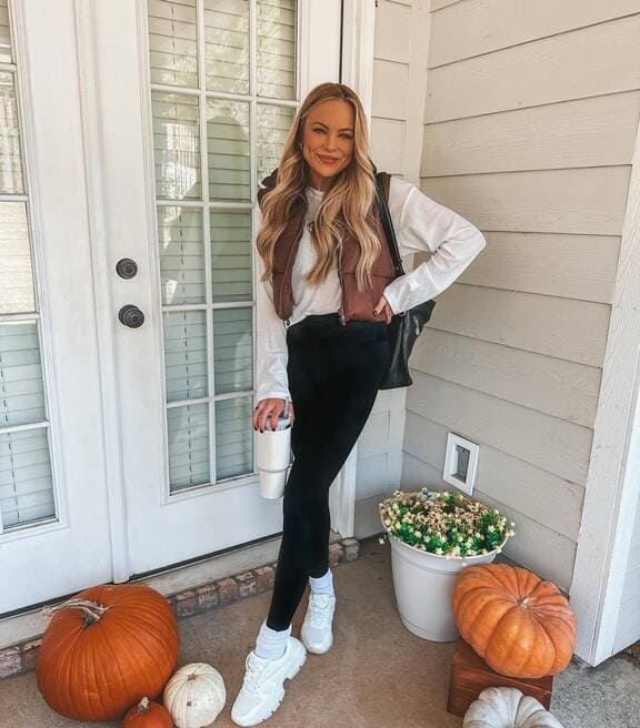 A woman is wearing a brown puffer vest, a white long-sleeve top, black leggings, white sneakers, and a tumbler.  