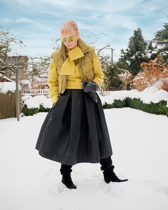 A woman wearing an olive puffer vest, yellow sweater, black midi skirt, and black boots in a snowy setting, accessorized with green sunglasses. 