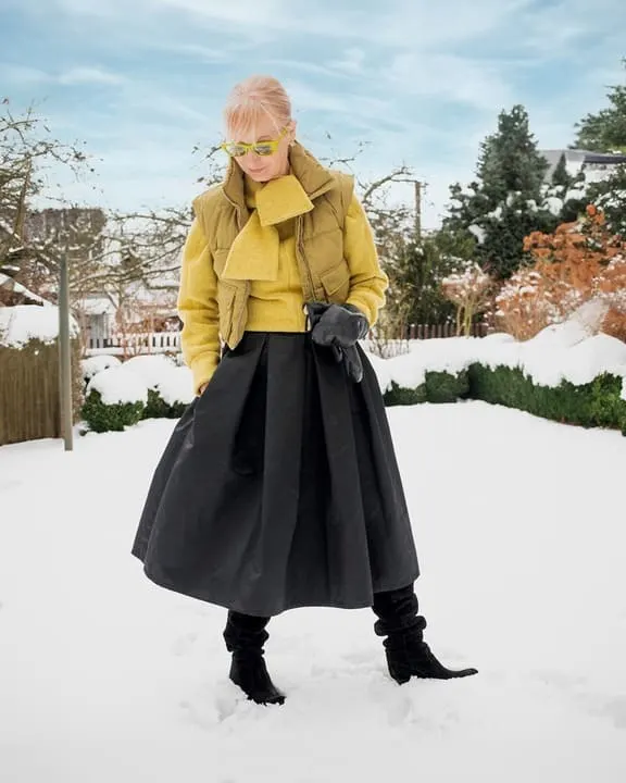 A woman wearing an olive puffer vest, yellow sweater, black midi skirt, and black boots in a snowy setting, accessorized with green sunglasses. 