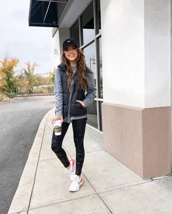 A woman wearing a black puffer vest, gray hoodie, black leggings, white sneakers, and a black baseball cap, holding a coffee cup. 