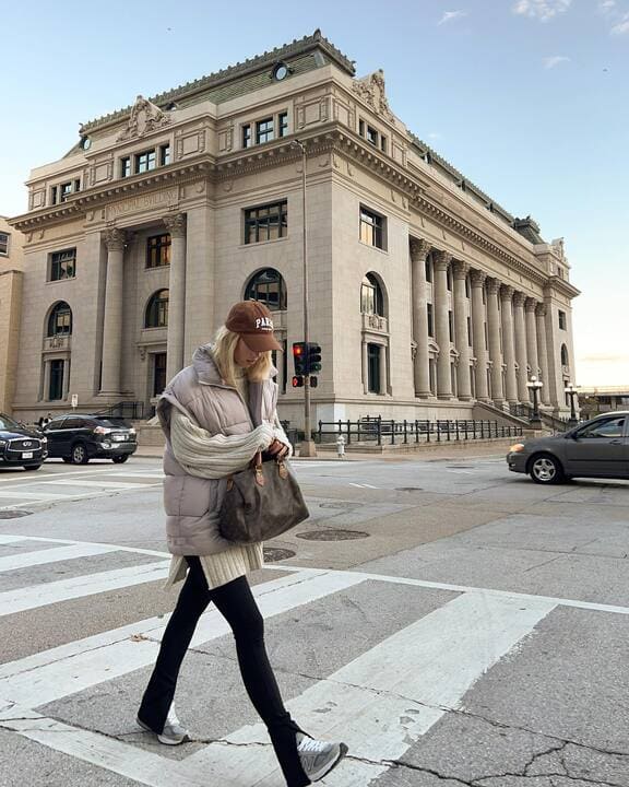 A woman wearing an oversized puffer vest, long knit sweater, black leggings, sneakers, and a brown cap while carrying a large handbag. 