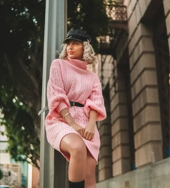 A woman is wearing a pink turtleneck sweater with black belt and a newsboy cap. 