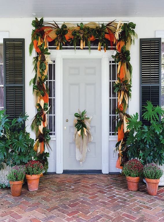 An elegant front porch decorated with a grand fall garland featuring magnolia leaves, burlap, and orange ribbons, creating a welcoming and seasonal entryway. 