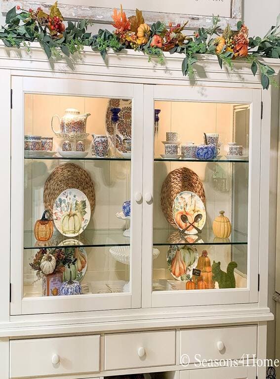 A festive fall hutch adorned with a vibrant garland of greenery, pumpkins, and colorful leaves, adding a lively seasonal touch. 