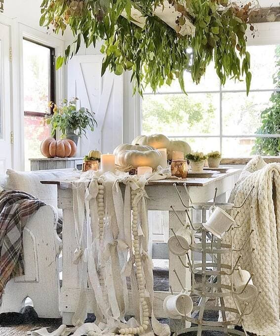 A serene fall tablescape featuring neutral-toned pumpkins, cozy candles, and cascading greenery overhead for a calming autumn atmosphere. 