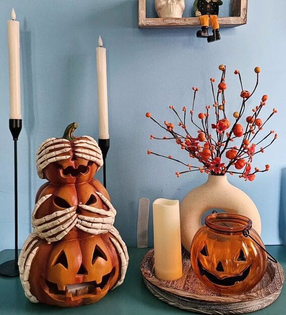 A quirky and charming vignette featuring stacked jack-o'-lanterns with skeleton hands, paired with a classic pumpkin lantern and vibrant orange berry branches. 