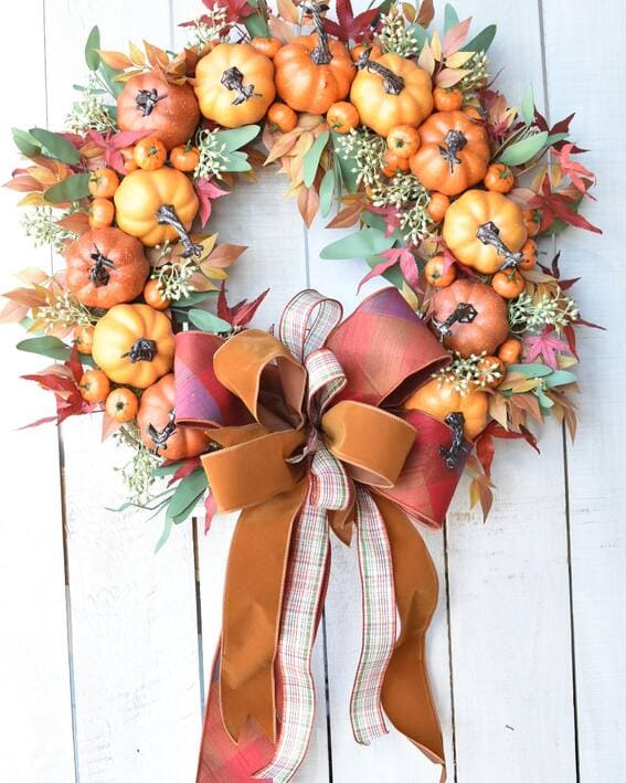 A fall wreath showcasing small pumpkins, vibrant autumn leaves, and a unique triple-layered bow in plaid, velvet, and satin.