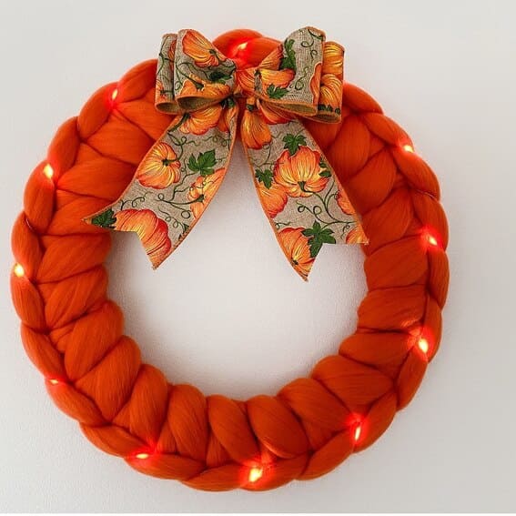 A bold and playful wreath made from braided wool in vibrant orange, accented with fairy lights and a pumpkin-themed bow. 