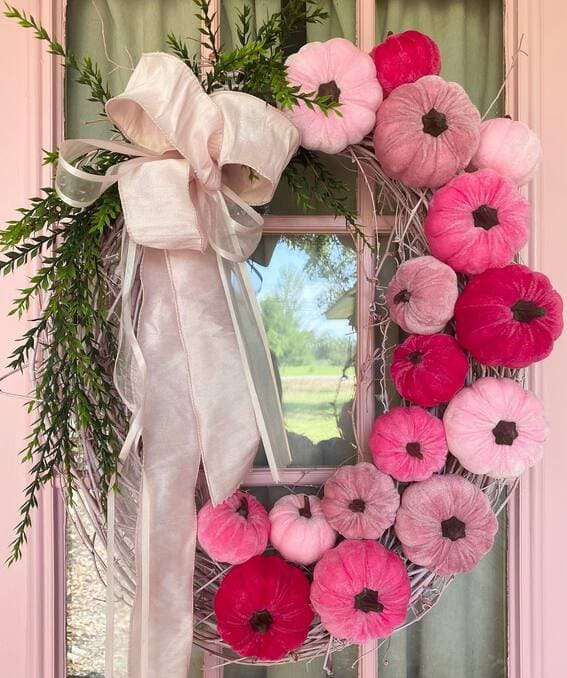 A whimsical wreath featuring plush velvet pumpkins in varying shades of pink, accented with soft greenery and an oversized satin bow. 