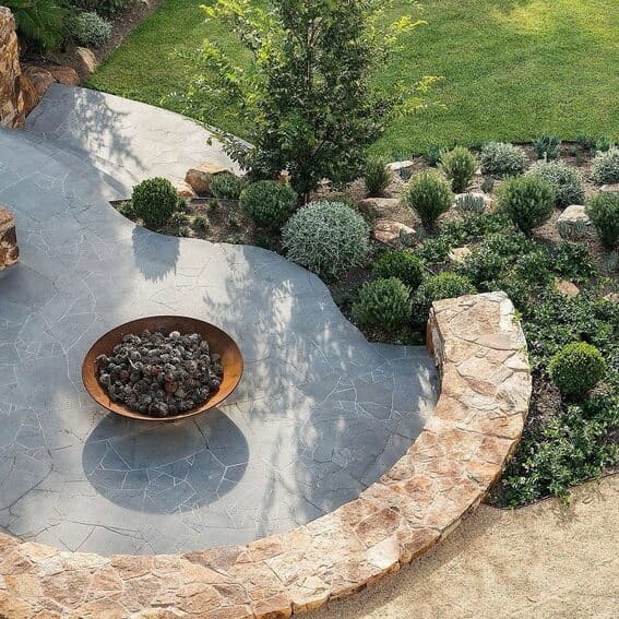 A curving stone patio with modern fire pit with lush vegetation. 