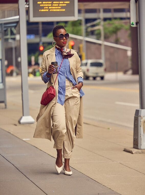 An elegant woman is styled in a blue-striped oversized shirt paired with a trench coat, tailored trousers, and bold accessories. 