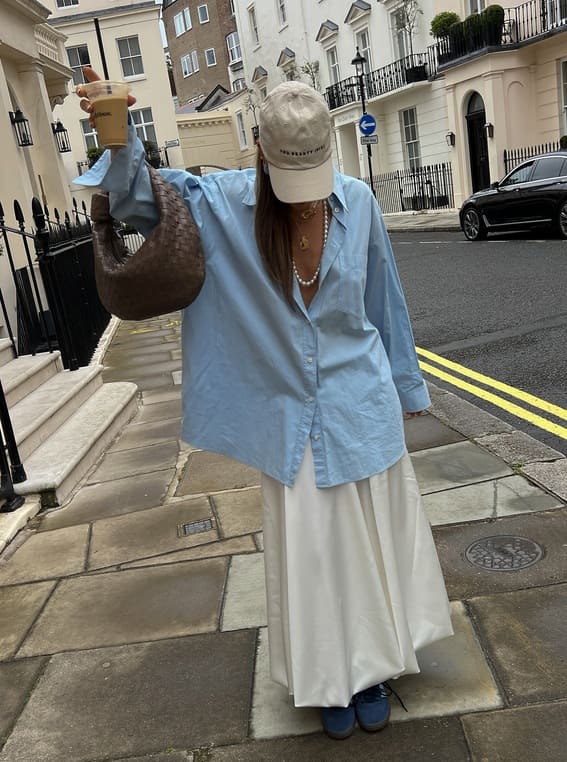 Effortlessly cool woman in an oversized blue shirt paired with a white maxi skirt and sneakers. 