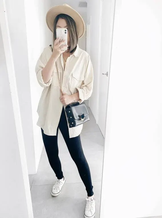 Chic woman in an oversized beige shirt paired with black leggings, white sneakers, and a fedora. 