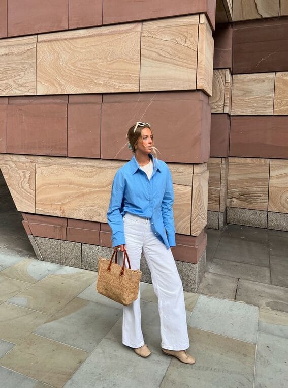 Chic woman in an oversized blue shirt paired with white wide-leg trousers and a woven bag. 