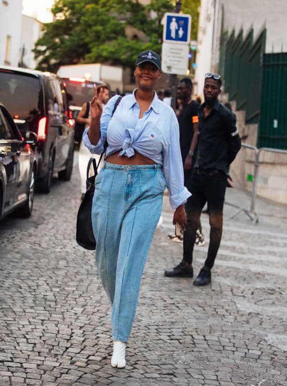 A confident woman in a knotted oversized blue shirt, high-waisted jeans, and white boots. 