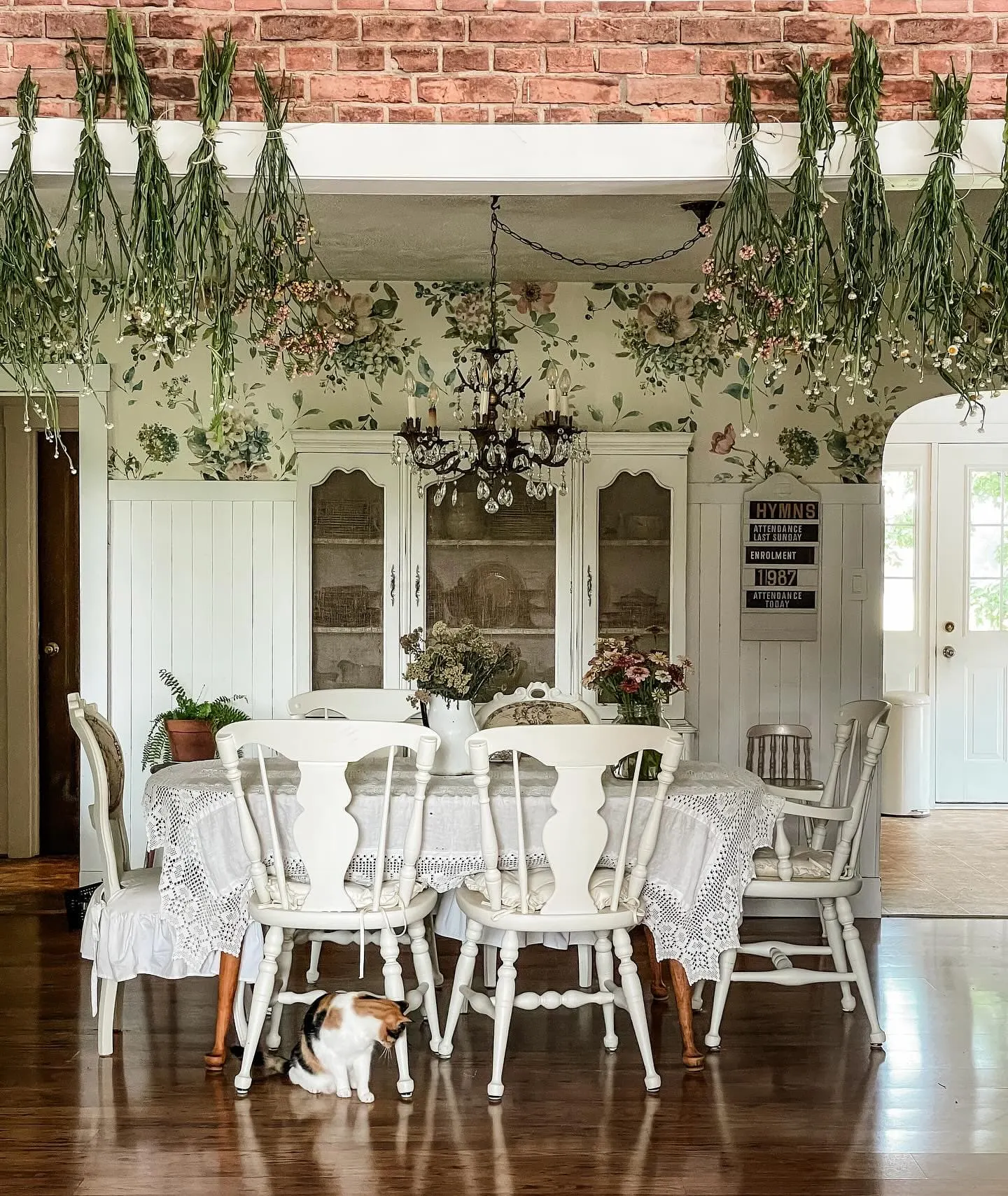 This image is an inspo for farmhouse dining, with floral wallpaper, hanging greenery, and a rustic chandelier that creates a warm, inviting atmosphere.Farmhouse dining room decor inspiration by @tracey_hiebert / Instagram